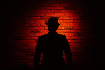 Silhouette of a man in a hat on a brick background. Noir, crime, detective.
