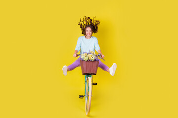 Full body size photo of charming funky young girl riding retro bike go highway fast carry flowers wind blow hair wear blue pullover violet pants isolated vibrant yellow color background