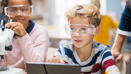 Elementary School Science Classroom: Boy Uses Digital Tablet Computer to Check Information on the Internet while Enthusiastic Cute Little Girl Uses Microscope.