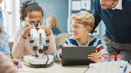 Elementary School Science Classroom: Cute Little Girl Looks Under Microscope, Boy Uses Digital Tablet Computer to Check Information on the Internet, while Enthusiastic Teacher Explains Lesson © Gorodenkoff