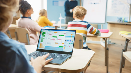 Elementary School Science Class: Over the Shoulder Little Boy Uses Laptop with Screen Showing Programming Software. Physics Teacher Explains Lesson to a Diverse Class full of Smart Kids