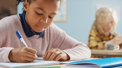 In Elementary School Classroom Brilliant Black Girl Writes in Exercise Notebook, Taking Test and Writing Exam. Junior Classroom with Group of Bright Children Working Diligently and Learning New Stuff