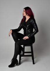 Full length portrait of  girl with red hair wearing black leather jacket, pants and boots. Sitting pose on a chair, isolated against a grey studio background.