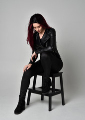 Full length portrait of  girl with red hair wearing black leather jacket, pants and boots. Sitting...