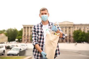 Male volunteer in protective mask and gloves with products on city street. Aid during coronavirus...