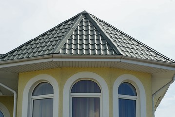 attic of a yellow house with white windows under a green tiled roof against the sky