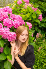 Obraz na płótnie Canvas Beautiful young blonde woman with flowers near face. Pink Hydrangea macrophylla blooming in summer in botanical garden