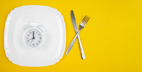 White alarm clock lying on a white plate with a fork and knife on a yellow background, banner, top view