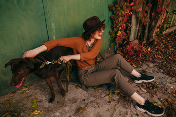 Happy lady in trendy vintage clothes sits on the ground in the yard of a country house and plays with a playful dog in the autumn day. Smiling owner having fun with dog on street near country house.