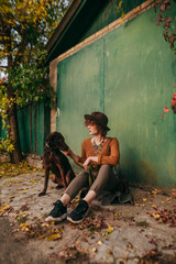 Fashion portrait of a lady sitting in the autumn on the ground on the background of the green gate of a country house and holding a dog on a leash, wearing vintage fashionable clothes. Vertical photo.