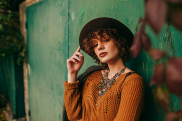 Autumn portrait of a curly-haired girl in a hat on a green grunge background and autumn leaves, looks into the camera and poses. Fashion portrait of lady on green wall background of country house.