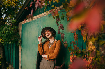 Smiling girl in fashionable clothes and hat stands in autumn day against the background of grunge green wall in country house,looking at camera and smiling. Fashion portrait of lady posing in village