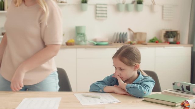 Medium shot of angry Caucasian woman is sitting at kitchen table, checking her daughter homework, scolding her then throwing pen on the table and going cooking meal. Girl is upset