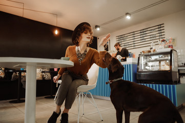 Lovely lady in trendy vintage clothes playing with dog sitting at coffee table with cup of coffee. Positive girl with a pet drooling in a cozy cafe. Animal friendly concept.