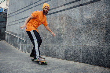 Obraz na płótnie Canvas Handsome young stylish hipster guy with beard in blank orange t-shirt riding on longboard.