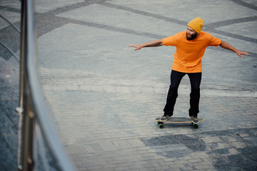 Handsome young stylish hipster guy with beard in blank orange t-shirt riding on longboard.