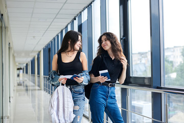Two young brunette girls, standing in light airport hallway with huge windows, wearing casual jeans clothes, holding international passports and boarding passes tickets. Girlfriends traveling by air.
