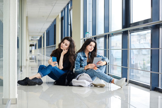 Two young brunette girls, sitting on floor in light airport hallway, with luggage behind, checking phone and tablet, wearing casual jeans clothes. Girlfriends, traveling by air, waiting for flight.