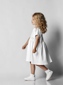 Cute smiling blonde curly kid girl in white casual dress and sneakers is walking, passing us over grey background