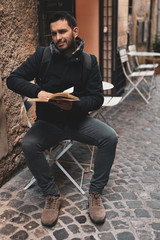 Handsome smiling man sitting on white chair outdoors and holding book in hands, man walking along the ancient streets, watching historical building exterior and learning history