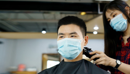 Fototapeta na wymiar Happy Asian man wearing a mask use haircut service in Barber shop. Woman hairdresser provide service mind. Hairdressing equipment is clean. End of quarantine and return to open salon hair work