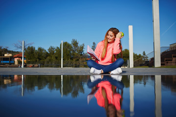 Portrait of female teenager reading absorbing book and eat green apple while sitting in the park, attractive young woman enjoy her holiday while read literature outdoors, student on the campus