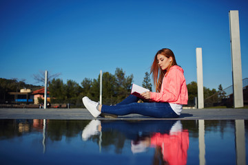 Portrait of female teenager reading absorbing book and eat green apple while sitting in the park, attractive young woman relaxing on her holiday while read literature outdoors, student on the campus