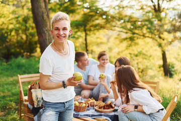 Positive guy with apple standing in front of people. Group of young people have vacation outdoors in the forest. Conception of weekend and friendship