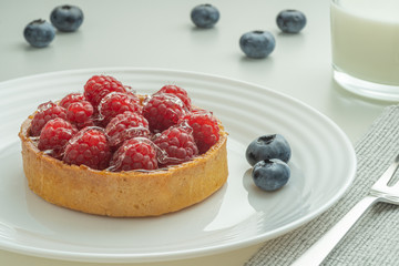 Raspberry tartlets served on a transparent plate with brown sugar. Glass of milk in the sun on a white background.