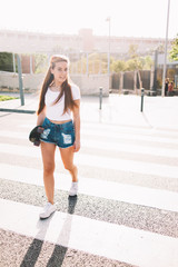 Full length portrait of smiling attractive hipster girl walking on the street with her skateboard during summer weekend, happy charming woman skater holding her penny board while cross the city road