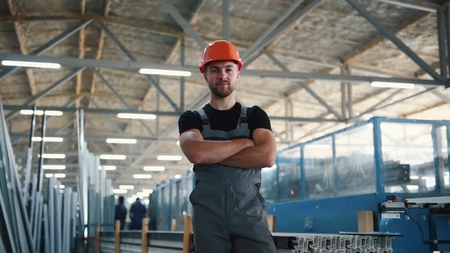 Worker in grey uniform and hard hat stands indoors in the factory with arms crossed.