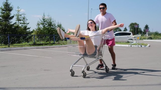 Friends are having fun in a shopping center parking lot on a bright day. energetic guy whirls in a circle cheerful girl in a basket enjoying free time slomo