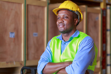 Young attractive happy African black man wearing yellow hard hat and safety vest standing and crossing his arms at warehouse, occupation job concept.