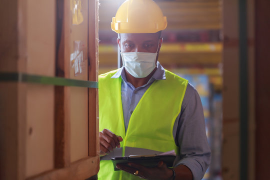 Young Black African Worker Wearing Protective Face Mask Working In Factory Warehouse. Black Man Checking Stock, New Normal After Covid 19 Pandemic Crisis. Logistic Industry Concept.