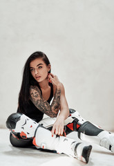 Fototapeta na wymiar Very beautiful looking brunette woman with cool tattoos in a black and red tracksuit, white boots and with a motorcycle helmet, posing in a white photo studio, sitting on the floor