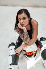 Fototapeta na wymiar Super beautiful looking brunette girl with tattoos in a black and red tracksuit, white and red boots and with a motorcycle helmet, posing in a white photo studio, sitting on the floor
