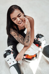 Fototapeta na wymiar Cute and funny looking brunette girl with tattoos in a black and red tracksuit, white boots and with a motorcycle helmet, posing in a white photo studio, sitting on the floor
