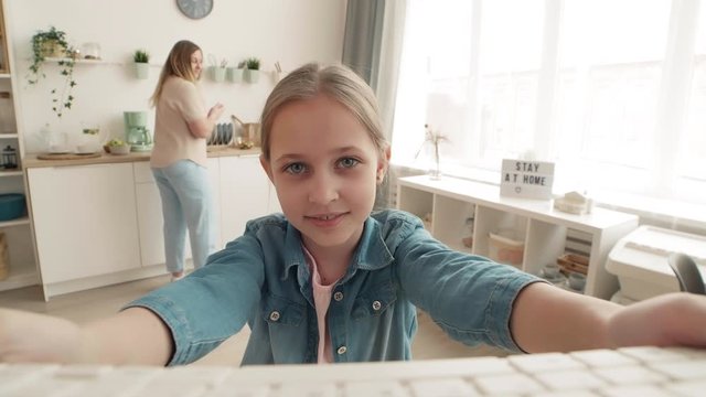 Caucasian girl holding laptop in her hands and having distance talk to friend while her mom cooking on background. Then woman coming to her daughter and greeting the interlocutor