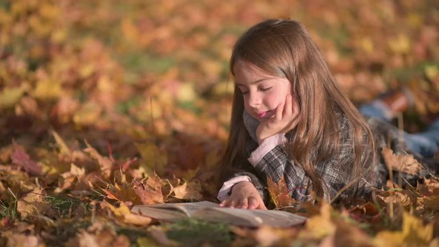 Sweet Girl in Polto Reads a Book Lying on Autumn Fallen Leaves. A Child Reads A Book in a Sunny Autumn Park.