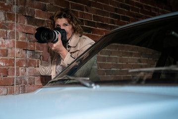 Eine Frau hält eine Kamera mit weißem Teleobjektiv vor das Gesicht. Die Journalistin fotografiert zwischen einer Mauer und einem hellblauen Auto.