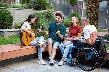 Drinks and songs. Group of friends taking a stroll on city's street in summer day. Handicapped man...