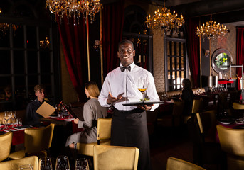 Handsome African-American waiter demonstrating tray of dish and wine Couple visitors on background