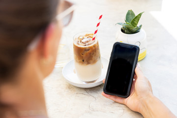 Girl enjoying an iced coffee while looking into her smartphone. Perspective from over the shoulder onto the smartphone.