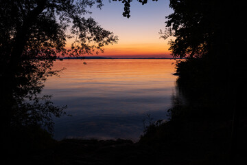 Colorful sunset at the lake Chiemsee in Bavaria, Germany. The picture is framed by the contour of plants and leafs. Water is very calm and reflects the colors.