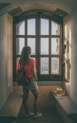 Pretty young woman standing by a window looking at the landscape.
Rear view of a pretty young woman standing by a 15th century window of a medieval castle. Tourism during the pandemic.