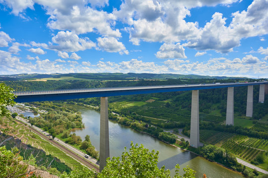 Beautiful Mosel Valley bridge made of blue steel and gray pillars, large trees in the background, blue sky with white gray clouds, railroad tracks can be seen. Germany.