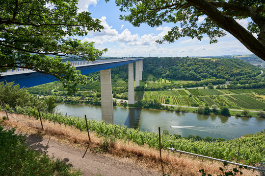 Beautiful Moselle Valley bridge made of blue steel and gray pillars, large trees in the foreground, blue sky with white-gray clouds, a narrow path leads past. Germany.