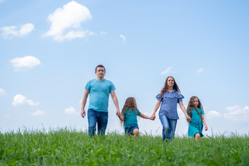 family dad, mom and two children walk on green grass against blue sky. Happy caucasian parents with two daughters holding hands.