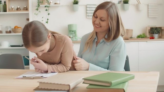 Medium shot of middle-aged Caucasian woman and blond-haired schoolgirl sitting at table at home, doing home task together, having fun and laughing