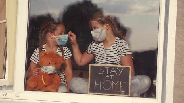 stay at home coronavirus. kids in medical mask in home with a toy teddy bear. kids holding a sign with the inscription stay at home. quarantine sitting by covid 19 the window. coronavirus concept